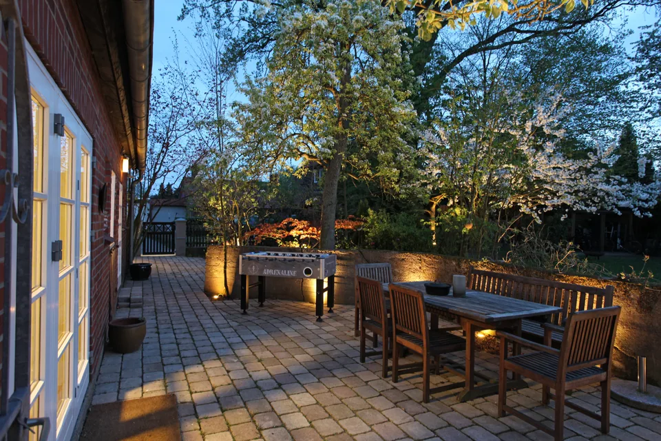 Beleuchtete Terrasse am Abend mit Holztisch, Sitzbänken und Tischkicker neben dem Garten.